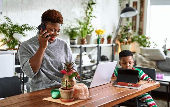 Smiling woman using laptop and talking on the phone whilst her young child watches a tablet nearby